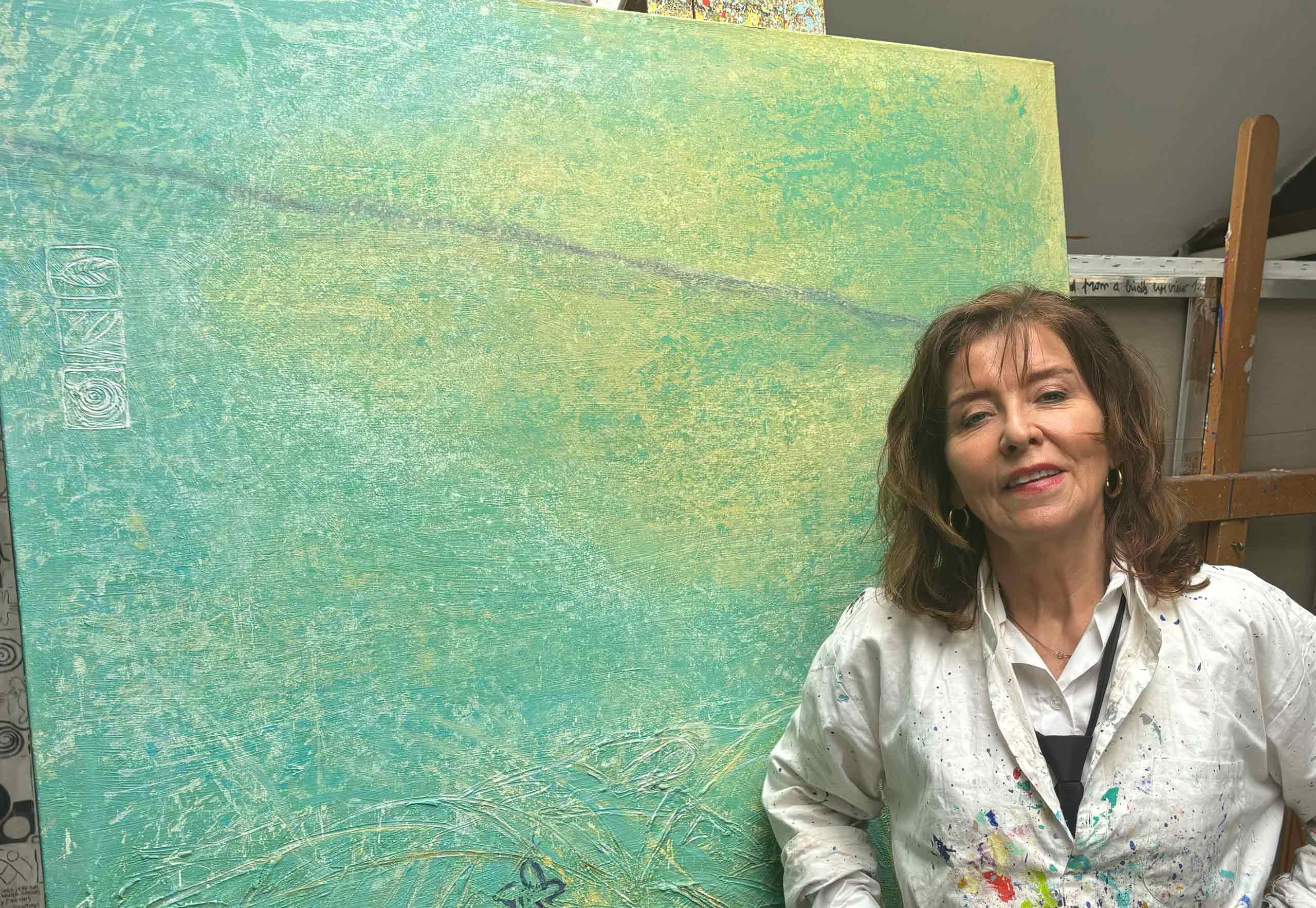 Merel-slide-2024-atelier_01 Kunstenaar Merel in haar schilderskiel voor een groot schilderij in groen- en turkooistinten in haar atelier
